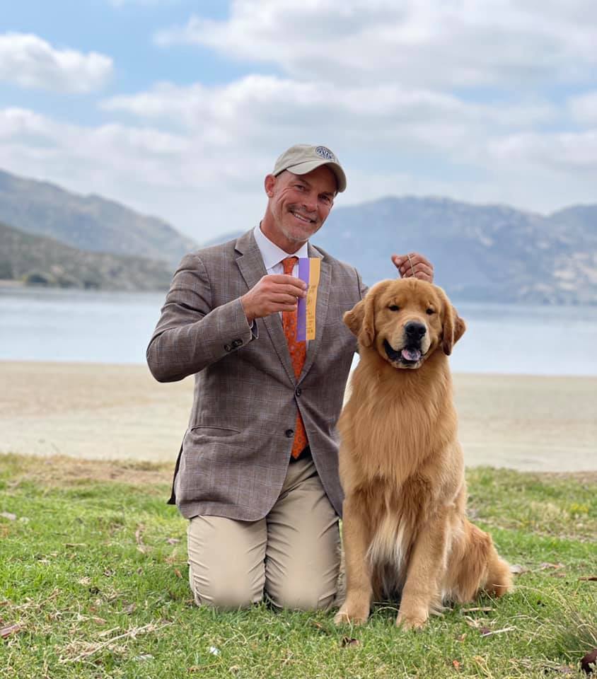 A man posing with a golden retriever in front of a lake.